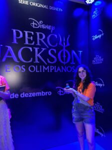 uma pessoa estava posando para a foto com um cupcake na mão, a luz é bem azul e há um backdrop escrito Percy Jackson atrás.
